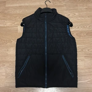 Svart quiltad dunväst med blått foder - Stilren svart dunväst med quiltad design och blått glansigt foder. Västen har två snedställda fickor med dragkedja och hög krage. Hör av vid intresse!