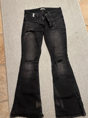 Svarta bootcut jeans Gina Tricot - Säljer ett par svarta bootcut jeans från Gina Tricot i storlek S. Jeansen har slitningar vid knäna och nertill, två stora framfickor och dubbla knappar i midjan. Materialet är klassisk jeans med en lite tvättad look och låg midja.
