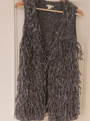 Grå fluffig väst från H&M XS - Säljer en grå fluffig väst från H&M i storlek XS. Västen har långa fransar framtill och är stickad i en mix av mörkgrå och ljusgrå toner. Perfekt för dig som vill ha en statement piece till din outfit.