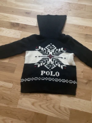 Svart polotröja Polo Ralph Lauren - Säljer en svart polotröja från Polo Ralph Lauren med stor vit snöflinga och Polo-text framtill. Tröjan har hög, ribbad polokrage med knappar, långa ärmar och stickat mönster i vitt och rött. Perfekt för kalla dagar och vintriga vibes.