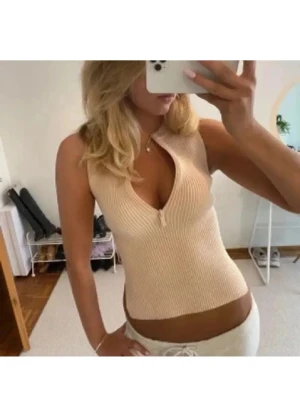 Beige ribbad ärmlös topp med dragkedja - Säljer en beige ribbad topp med croppad passform och liten dragkedja framtill. Toppen är ärmlös och har en snygg v-ringning. Perfekt för varma dagar och enkel att styla med jeans eller kjol.