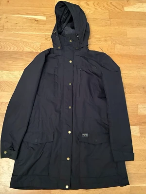 Svart parkas från Peak Performance - Säljer en svart parkas från Peak Performance med huva, dragkedja och tryckknappar. Jackan har flera praktiska fickor, justerbar midja och är tillverkad i slitstark polyester. Perfekt för kyliga dagar och outdoor-äventyr.