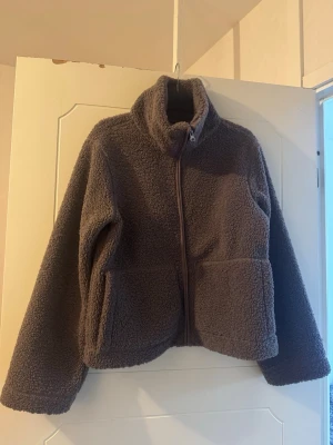 Brun lila teddyjacka - Mysig brun teddyjacka från i storlek S. Jackan har hög krage, dragkedja framtill och två stora fickor. Perfekt för kyliga dagar och ger en avslappnad och trendig look. Materialet är fluffigt och mjukt, vilket gör den superbekväm att bära.