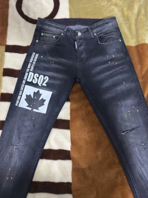 Svarta DSQUARED2 slim jeans med tryck - Säljer ett par svarta slim fit jeans från DSQUARED2 i storlek 44. Jeansen har coolt vitt tryck med lönnlöv och text på vänster ben, samt slitningar och färgstänk för en edgy look. Klassisk femficksmodell med knappgylf och röda sömdetaljer på insidan.