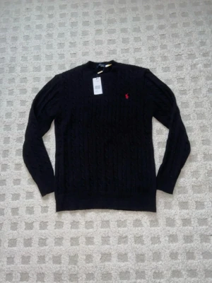 Svart kabelstickad Ralph Lauren tröja - Kabelstickad svart tröja från Polo Ralph Lauren med klassisk röd broderad logga på bröstet. Tröjan har rund halsringning och långa ärmar. Tillverkad i mjuk ull som känns riktigt skön mot huden. Perfekt för dig som vill ha en stilren och tidlös look. QR-koden samt serienumret funkar!