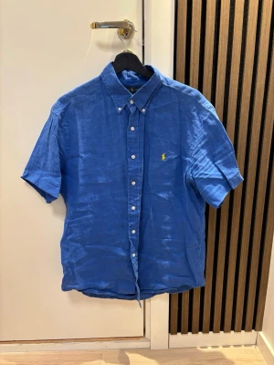 Blå linneskjorta från Ralph Lauren - Kortärmad blå skjorta från Ralph Lauren i svalt linne. Klassisk button-down krage, vita knappar framtill och gul broderad logga på bröstet. Perfekt för varma dagar och enkel att styla till jeans eller shorts. Kom dm för eventuella frågor🧐pris kan diskuteras 💯😇