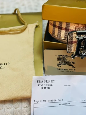 Rutig Burberry bälte med B-spänne - Säljer ett klassiskt Burberry bälte med ikoniskt rutigt mönster i beige, brun och röd. Bältet har ett stort metallspänne med Burberrys B-logga och är gjort i skinn. Kommer med originalförpackning och dustbag.