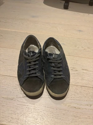 Svarta blåa Golden Goose sneakers i mocka - Säljer ett par svarta Golden Goose Superstar sneakers med klassisk stjärndetalj på sidan. Skorna har snörning, vit sula och är gjorda i mocka. Perfekta för dig som gillar en avslappnad och trendig stil.