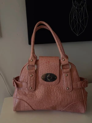 Rosa axelväska i krokopräglat skinn - Säljer en snygg rosa axelväska i krokopräglat skinnimitation med silverfärgade metalldetaljer och vridlås framtill. Väskan har dubbla handtag, justerbara spännen på sidorna och flera fack inuti för smart förvaring. Perfekt för dig som vill ha en unik och trendig accessoar.
