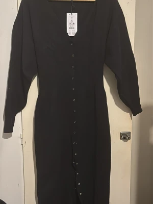 Svart långklänning från Gina Tricot - Svart långklänning från Gina Tricot med v-ringning och långa ärmar. Klänningen har knappar hela vägen framtill och är tillverkad i ett mjukt material med 50% LENZING™ ECOVERO™ viskos. Enkel och stilren design som passar till många tillfällen.