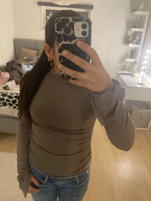 Brun långärmad polotopp - Säljer en brun långärmad polotopp med tight passform. Toppen har långa ärmar med lite extra tyg vid ärmsluten och är gjord i ett stretchigt material som sitter snyggt mot kroppen. Perfekt att styla med jeans eller kjol.