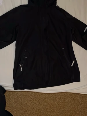 Svart vindjacka från Helly Hansen - Säljer en svart vindjacka från Helly Hansen med huva och vita dragkedjor. Jackan har meshfoder på insidan, två fickor med dragkedja och justerbar huva. Perfekt för blåsiga dagar och outdoor-äventyr.