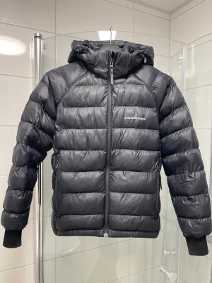 Svart dunjacka från Peak Performance - Säljer en svart dunjacka från Peak Performance med huva och dragkedja framtill. Jackan har ribbade muddar vid ärmsluten och två sidofickor med dragkedja. Perfekt för kalla dagar och riktigt snygg till alla outfits.