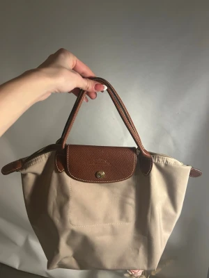 Beige handväska från Longchamp - Säljer en klassisk beige handväska från Longchamp med bruna skinnhandtag och lock. Väskan har guldfärgad knapp och dragkedja upptill. Perfekt storlek för vardagens essentials och enkel att matcha till olika outfits.