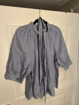 Kortärmad blus från Holly & Whyte - Säljer denna charmiga blus från Holly & Whyte med klassiskt blåvitt randigt mönster. Har ett matchande skärp/band att knyta i midjan för en smickrande silhuett. Perfekt för en stilfull och avslappnad look. Ett måste i garderoben! Passar M/L.