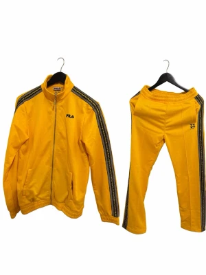 Gula FILA träningskläder set - Snyggt gult träningsset från FILA med svarta detaljer längs ärmar och ben. Jackan har dragkedja och ståkrage, byxorna har resår i midjan och båda delarna har logga.