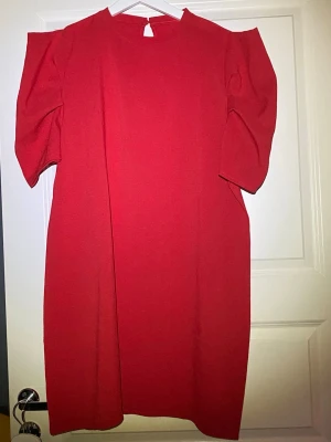 Röd kortklänning med puffärm - Säljer en röd kortklänning med puffiga kortärmar och rund hals. Klänningen har en enkel knappdetalj i nacken och är gjord i ett mjukt tyg som känns bekvämt. Perfekt för dig som vill ha en stilren men ändå statement look.