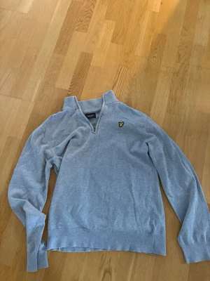 Grå half zip tröja Lyle & Scott - Snygg ljusgrå tröja från Lyle & Scott med klassisk gul logga på bröstet. Tröjan har half zip-dragkedja vid halsen och är långärmad. Perfekt till chill vardagsstil och enkel att matcha med jeans eller sweatpants.