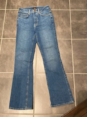 Blå Lee Breese Bootcut jeans W27 L31 - Säljer ett par klassiska blå bootcut jeans från Lee, modell Breese Boot. Jeansen har fem fickor, normal passform och snyggt utsvängda ben. Tillverkade i slitstarkt denim med kontrastsömmar och dragkedjegylf. Perfekta för dig som gillar retrovibbar och tidlös stil.