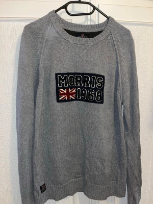 Grå stickad tröja från Morris - Säljer en grå stickad tröja från Morris med broderad patch på bröstet där det står 'Morris 1968' och en brittisk flagga. Tröjan har rund halsringning, ribbade muddar och långa ärmar med diskret kabelstickad detalj längs axlarna. Perfekt för dig som gillar klassisk men sportig stil.
