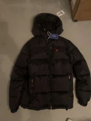 Svart pufferjacka från Polo Ralph Lauren - Säljer en svart pufferjacka från Polo Ralph Lauren med huva och dragkedja. Jackan har det klassiska röda Polo-logot på bröstet och två fickor med dragkedja. Perfekt för kalla dagar och riktigt snygg till streetwear-stilen.