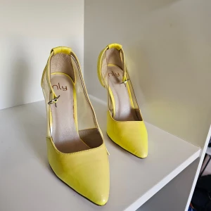 Gula pumps med transparent detalj från Nly - Snygga gula pumps från Nly med spetsig tå och hög klack. Skorna har transparenta sidor i syntetmaterial och en smal rem runt vristen med spänne. Perfekta för dig som vill sticka ut med färg och cool design.