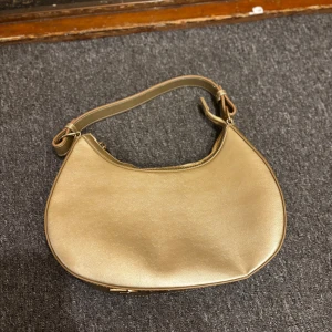 Guldig axelväska från RBYR - Snygg axelväska i guldigt syntetmaterial med stilren bågform och bred axelrem med RBYR-logga i metallic. Väskan har dragkedja upptill och minimalistisk design som sticker ut. Perfekt för dig som vill ha en trendig accessoar med lite extra bling.