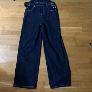 Mörkblå denim hängselbyxor wide - Snygga mörkblå denim hängselbyxor med breda ben och kontrastsömmar. Klassisk design med justerbara axelband, fickor fram och bak samt coola metallknappar i sidorna. Perfekta för en avslappnad och trendig look.