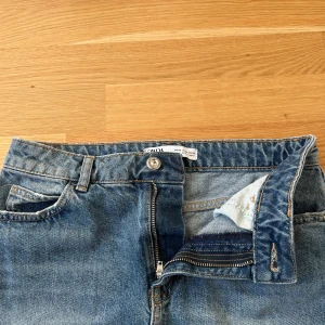 Blå jeanskjol från ZARA med rå kant - Snygg blå jeanskjol från ZARA med klassisk femficksdesign, dragkedja och knapp framtill. Kjolen har rå, fransig kant nedtill och bälteshällor. Midjemåttet är 33,5cm och den är 38cm lång. 