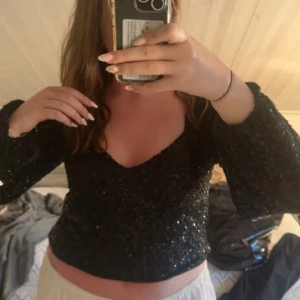 Svart paljettopp från Gina Tricot - Säljer en svart glittrig topp från Gina Tricot med paljetter över hela. Toppen har lång ärm, croppad passform och en snygg v-ringning. Knappt använd!