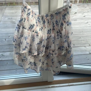 Blommig volangkjol från Vero Moda - Supersöt kort kjol från Vero Moda med blommigt mönster i blått och ljusrosa på vit botten. Kjolen har smock i midjan och flera lager volanger som ger en flowy känsla. Perfekt för sommarens alla tillfällen!