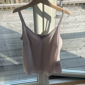 Ljusrosa linnetopp från All Saints - Supersnygg ljusrosa linnetopp från All Saints med smala axelband och v-ringning. Toppen har en stilren och enkel design i ett slätt, mjukt material som ger en lyxig känsla. Perfekt att styla med jeans eller kjol för en fräsch look.