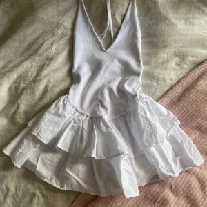 Vit omlottklänning med volanger från Zara - Supersöt vit kortklänning från Zara med omlott-design och djup v-ringning. Klänningen har tunna axelband och två lager volanger i kjolen som ger extra flärd. Perfekt för sommarens alla tillfällen, med en lätt och luftig känsla.