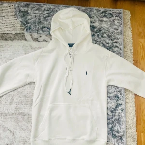 Vit hoodie från Polo Ralph Lauren - Snygg vit hoodie från Polo Ralph Lauren med klassisk logga broderad på bröstet. Tröjan har huva med snörning, känguruficka och ribbade muddar. Perfekt för en clean och stilren look. Tillverkad i mjuk bomull för extra komfort.