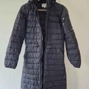 Svart lång lättvikts dunjacka från CRW - Lång svart lättvikts dunjacka från CRW med horisontella sömmar, huva och dragkedja hela vägen. Jackan har en ficka med dragkedja på ärmen och två sidofickor. Perfekt för kalla dagar och riktigt snygg till vardagslooken.