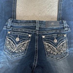 Miss Me blå jeans med paljettdetaljer - Snygga blå jeans från Miss Me med vita kontrastsömmar och unika bakfickor dekorerade med paljetter och broderier. Modellen har midwaist och bootcut, med coola detaljer som glittrande knappar. Perfekta för dig som gillar statement-jeans.