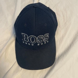 Mörkblå keps från Hugo Boss - Snygg mörkblå keps från Hugo Boss med stor vit broderad logga framtill. Klassisk böjd skärm och ventilerande mesh-tyg. Perfekt accessoar för en sportig och stilren look. Justerbar baktill för bästa passform.