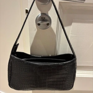 Svart axelväska med krokoprägling - Snygg svart axelväska med krokopräglat mönster och dragkedja upptill. Perfekt storlek för det viktigaste och har en stilren, rektangulär form. Väskan har ett trendigt och minimalistiskt uttryck som passar till många outfits.