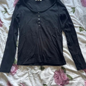 Svart långärmad topp från H&M - En svart långärmad topp från H&M i ribbad bomull. Modellen har rund halsringning och knappar framtill som ger en enkel men snygg look. Perfekt att matcha med jeans eller kjol för en chill stil.