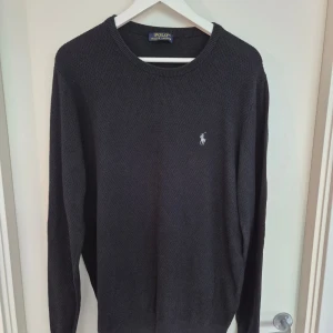 Svart stickad tröja från Polo Ralph Lauren - Klassisk svart stickad tröja från Polo Ralph Lauren i storlek XXL. Tröjan har rund halsringning, lång ärm och den ikoniska broderade loggan på bröstet. Perfekt för dig som gillar en enkel och stilren look.