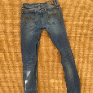 Nudie Jeans - Säljer ett par blå skinny jeans från Nudie Jeans i storlek 32/32. Jeansen har klassisk femficksdesign, snygga slitningar och Nudie-loggan broderad på bakfickorna. De är i stretchigt denim med orangea sömmar och har en smal passform. Slim fit, bra skick.