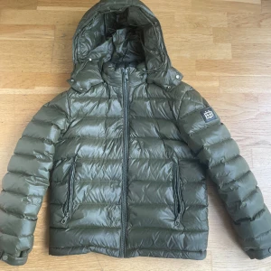 Bvaldi - Pufferjacka - Snygg olivgrön pufferjacka från Bvaldi med huva och två dragkedjeförsedda fickor. Jackan har ett stilrent emblem på ärmen och är tillverkad i 100% polyamid. Perfekt för kalla dagar när du vill hålla stilen och värmen. Jackan är i nyskick och endast använda några gånger. Säljer pga att den är för liten. Nypris 3k