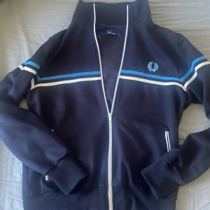 Blå track jacket från Fred Perry Storlek M - Säljer en snygg blå track jacket från Fred Perry med vita och blå ränder över bröstet och ärmarna. Jackan har dragkedja framtill, ribbade muddar och klassisk Fred Perry-logga broderad på bröstet. Perfekt för en sportig och stilren look. I storlek M