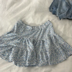 Blommig volangkjol från 157  - Supersöt kort volangkjol i ljusblått från 157  med vita och svarta blommor. Kjolen har resår i midjan och flera lager volanger som ger en flowy känsla. Perfekt för vår och sommar när du vill ha en chill och söt look. 