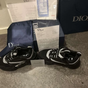 Svarta Dior sneakers med silvriga detaljer - Snygga svarta Dior sneakers med silvriga detaljer och meshpaneler. Skorna har rund tå, snörning och en chunky sula. Märkeslogga på insidan och coola kontrasterande partier i syntet och skinn. Perfekta för dig som vill sticka ut med exklusiva och trendiga sneakers.