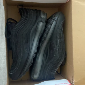 Nike Air Max 97 Triple Black 38.5 (DAM) - Säljer ett par Nike Air Max 97 i helsvart färg. Skorna har den ikoniska vågiga designen, synlig Air-sula och snörning. Ovandelen är i syntet och mesh för en sportig look. Perfekta för dig som gillar streetstyle och vill ha en clean sneaker. Nästan oanvända. Storlek 38,5.