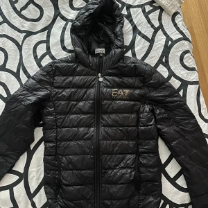 Svart pufferjacka från EA7 Emporio Armani - Snygg, sportig och supervarm! Denna svarta pufferjacka från EA7 Emporio Armani har en quiltad design, huva och en elegant guldfärgad logga på bröstet – en riktig statement-jacka. Med dragkedja framtill och två praktiska sidofickor är den både stilren och funktionell. Perfekt för kyliga dagar, och garanterat att höja din look!
