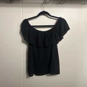 Svart offshoulder topp från H&M XS - Svart offshoulder topp från H&M i storlek XS. Toppen har bred volang runt hela axelpartiet och är ärmlös, vilket ger en snygg och luftig look. Perfekt för dig som gillar en enkel men trendig stil.