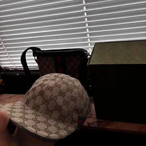 Gucci keps/gucci väska combo  - Snygg keps från Gucci i beige med klassiskt GG-monogram över hela kepsen. Den har en böjd skärm och detaljer med grönt och rött band på sidan. Materialet är bomull och polyester, vilket gör den både bekväm och stilren. Perfekt för dig som vill ha en lyxig streetwear-look.