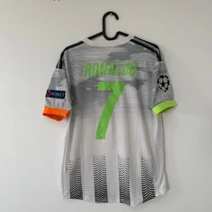 Juventus Ronaldo #7 Adidas fotbollströja M - Juventus matchtröja med Ronaldo #7 på ryggen, Adidas-märke och Jeep-logga i neon-grönt. Svartvita ränder med prickigt mönster, Champions League och Respect-patch på ärmarna, ena ärmen har neon-grön kant och den andra orange. Tillverkad i lätt polyester.
